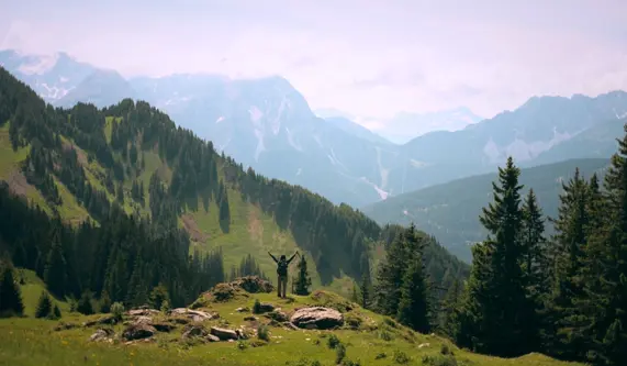 A person enjoys the view from a hill, with towering mountains stretching into the distance.